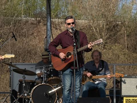 Full band on an outdoor covered stage with spring trees in background